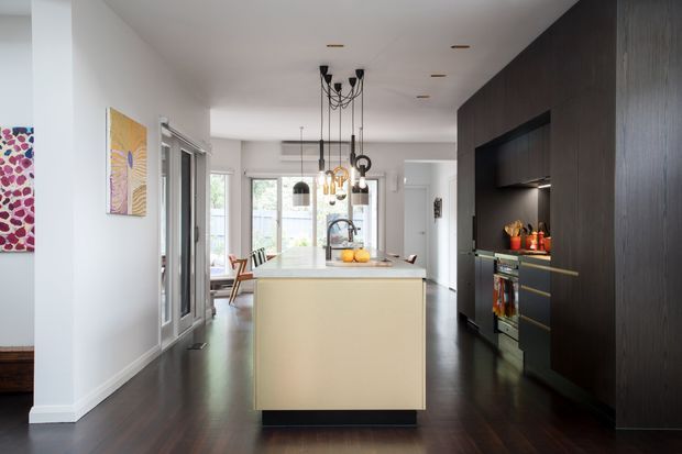 A contemporary open-plan kitchen viewed from the adjoining living area, featuring a large central island, dark timber cabinetry along the right wall, integrated appliances, pendant lighting, white walls, and dark timber flooring. A dining and living area is visible beyond the kitchen, with colourful artwork adding accents.
