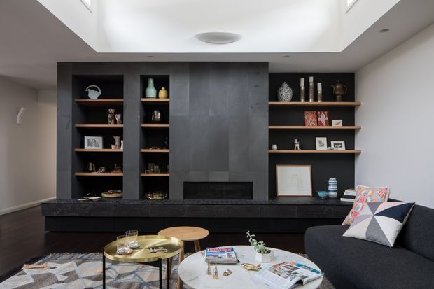 Contemporary living room featuring a dark built-in entertainment and fireplace wall with open timber display shelving, a low dark stone hearth, and a recessed gas fireplace. A charcoal sofa, patterned cushions, layered coffee tables, and a geometric rug furnish the room. A large recessed ceiling light well provides bright overhead illumination.