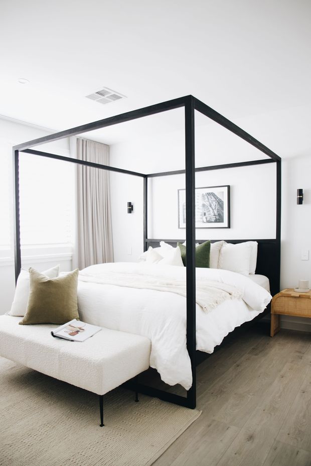 Bright, refined bedroom featuring a black four-poster canopy bed with crisp white bedding, layered neutral cushions, a textured upholstered bench at the foot, pale timber flooring, and a large light rug. White walls, full-height curtains, minimalist black wall sconces, framed monochrome artwork, and a woven bedside table create a calm contemporary look.