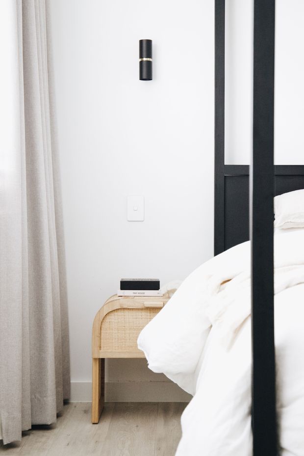 A softly lit bedroom corner featuring a black metal four-poster bed with white bedding, a pale timber and woven-cane bedside table, light timber flooring, sheer beige curtains, and a minimal white wall with a black cylindrical wall light.