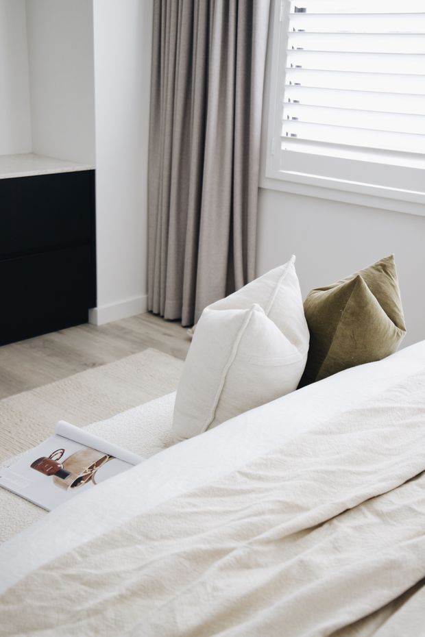 Bright, softly styled bedroom corner featuring a neatly made bed with white textured linen, white and olive-green cushions, pale timber-look flooring, a light rug, full-length taupe curtains, white plantation shutters, and a dark built-in cabinet with a pale stone or light benchtop.