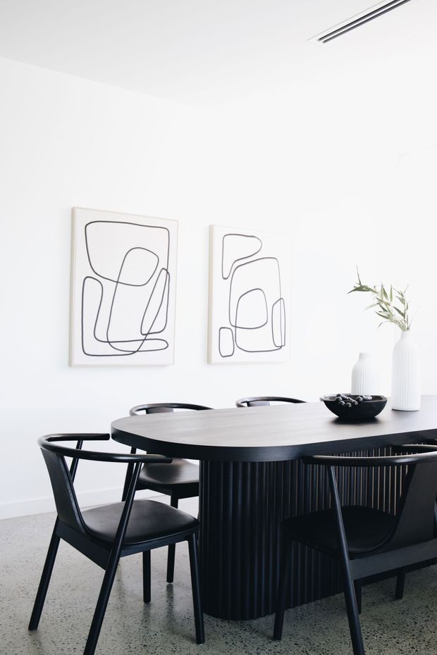 A bright, minimalist dining area with a large oval dark timber dining table, black sculptural dining chairs, and a vertically fluted black table base. White walls feature two abstract line artworks, while a white vase with greenery and a black bowl provide restrained styling. The room has a polished light terrazzo floor and a slim linear ceiling air vent or light detail.