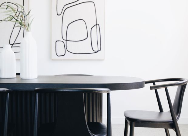 Minimalist dining area with a dark oval dining table and black chairs against a bright white wall. Abstract black-line artwork, sculptural white vases, and subtle plant foliage add restrained decorative detail.