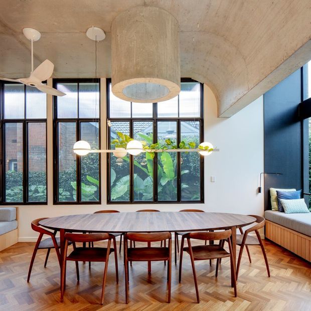 A bright, carefully detailed dining room with a large oval timber dining table and mid-century-style wooden chairs. Floor-to-ceiling black-framed windows overlook leafy planting, while a built-in window seat runs along the right wall. The room features a striking exposed concrete ceiling with a cylindrical concrete pendant enclosure, a sculptural linear dining light, ceiling fans, and warm herringbone timber flooring.