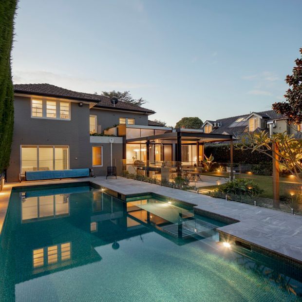 A dusk exterior view of a contemporary suburban residence with a large swimming pool in the foreground, paved pool deck, landscaped garden, and a covered outdoor entertaining pavilion with glazed walls and warm interior lighting. The two-storey home has dark painted masonry walls, tiled pitched roofs, large windows, and a sheltered outdoor dining or lounge area.