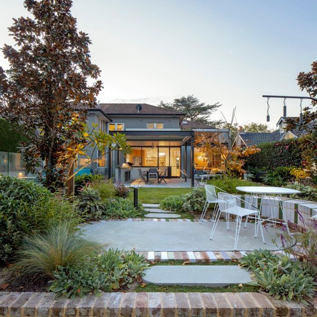 A landscaped residential courtyard and rear garden viewed from outside, with a contemporary two-storey grey house beyond. The garden features a broad concrete terrace, white outdoor dining furniture, stepping-stone paths, layered drought-tolerant planting, mature trees, hedges, and subtle outdoor lighting. The house has dark-framed glazing, a covered patio connection, and a tiled pitched roof.