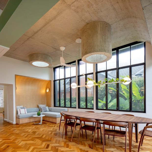 A bright open-plan dining and living area with a long timber dining table and mid-century style wooden chairs, a built-in light upholstered sofa, expansive black-framed windows, and a distinctive timber-lined ceiling with cylindrical concrete pendant light housings. The floor is laid in a warm herringbone timber pattern, and leafy greenery is visible outside the glazing.