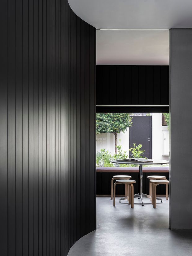 A minimalist dark-toned interior looking through a curved opening into a compact dining or breakfast area. Vertical black wall panelling frames the view, with a smooth grey ceiling and polished grey floor. The adjoining space contains a slim dark table, light timber stools, and a large opening to a planted courtyard, creating a strong indoor–outdoor connection.