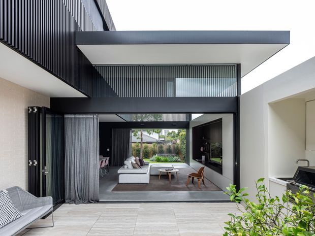 A contemporary indoor-outdoor living space viewed from a bright tiled terrace or courtyard. The open-plan living room features large black-framed glazing, a white sectional sofa, timber lounge chairs, a low round coffee table, dark built-in joinery and full-height curtains. Strong vertical screening and a projecting flat roof create a crisp modern architectural composition. A compact kitchen or utility area is visible at the right edge, while greenery appears in the foreground and garden beyond.