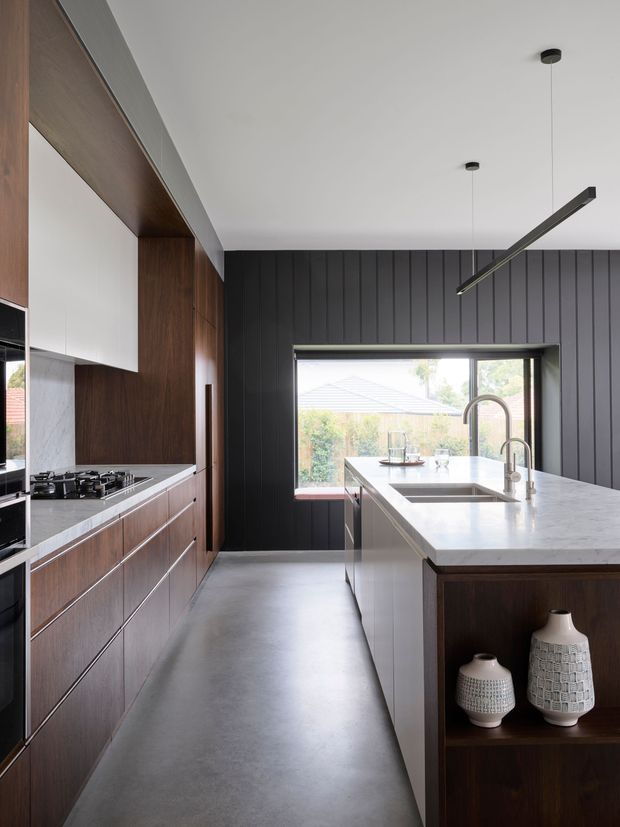 A modern galley-style kitchen with a large central island, dark vertical-panel feature walls, and warm dark timber cabinetry. The island has a pale stone benchtop, integrated sink, and curved brushed-metal tapware. A gas cooktop and pale marble-look splashback sit along the left wall, while slim linear pendant lights hang from a white ceiling. A window above the island overlooks greenery and neighbouring rooftops. Decorative ceramic vessels are displayed in open timber shelving at the end of the island.