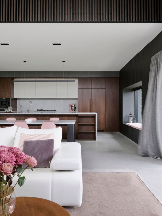 A sophisticated open-plan living and kitchen interior viewed from the living area. The room features a white sectional sofa, pale rug, pink and mauve cushions, and a round timber coffee table in the foreground. Behind it is a streamlined kitchen with white upper cabinetry, a marble-look splashback, dark timber cabinetry, a long island with seating, and integrated appliances. Dark vertical timber battens line the ceiling edge and right-hand wall, complemented by grey flooring, recessed lighting, and floor-to-ceiling curtains. The overall look is refined, contemporary, and warm with strong dark-and-light contrast.