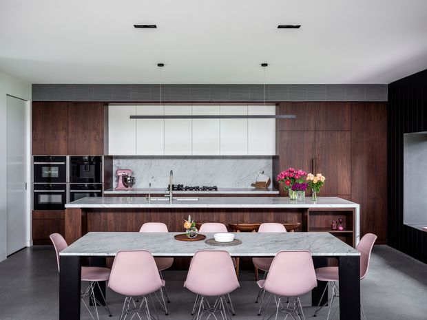Contemporary kitchen and dining area with a large marble-look island and dining table, dark timber cabinetry, integrated black appliances, pale pink dining chairs, and a black ribbed feature wall. Two linear pendant lights and recessed ceiling lighting provide a clean, refined fit-out.