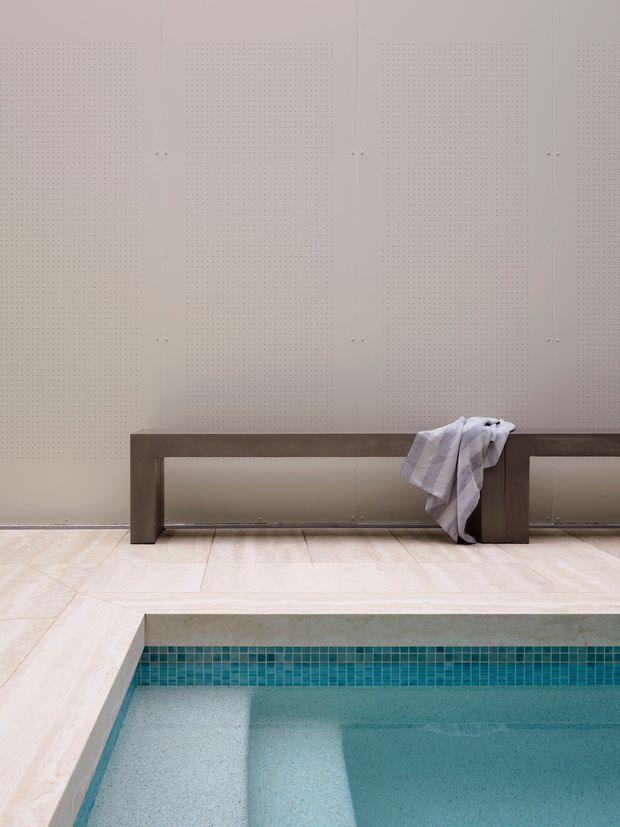 A minimalist indoor pool area with a pale tiled surround, a recessed turquoise tiled pool, and a long dark timber or stained-wood bench against a wall of light perforated cabinetry or acoustic panelling. A light towel is draped over the bench, adding a subtle lived-in detail. The restrained neutral palette and clean lines create a calm, spa-like atmosphere.