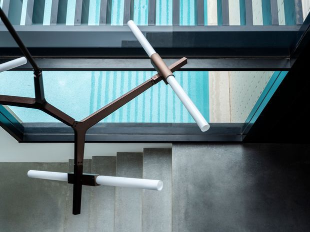 Upward-looking architectural interior view beneath a glazed roof or skylight, with dark structural framing, translucent turquoise glazing, a sculptural branching pendant light with white tubular arms, and a concrete or stone stair rising along the lower portion of the image. The space has a dramatic, minimalist contemporary character.