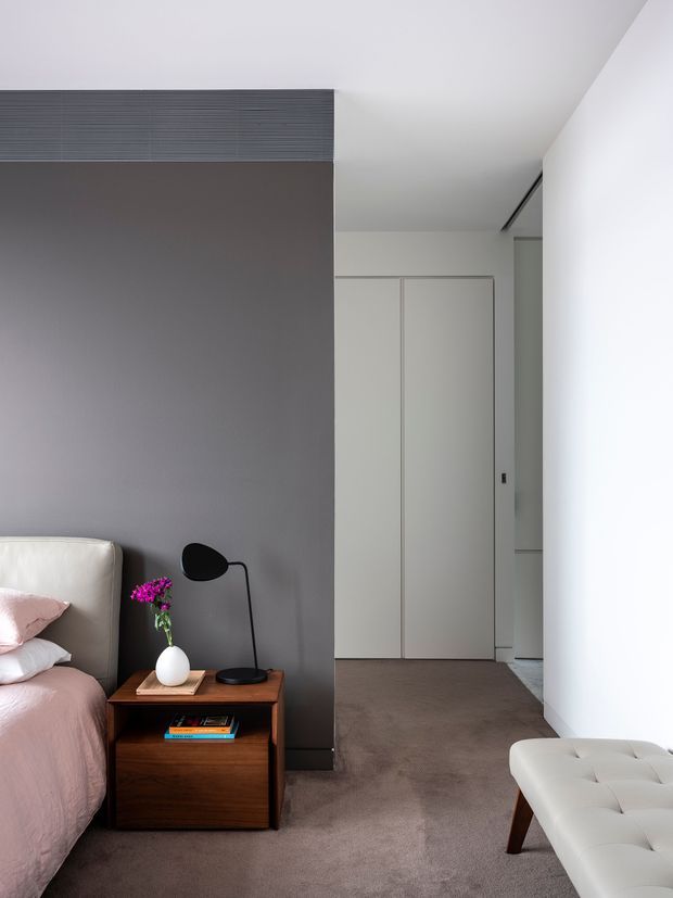 A minimalist bedroom with a partially visible bed dressed in blush pink bedding, a dark grey feature wall, warm timber bedside table, black table lamp, white ceramic ornament, and a light upholstered bench. Built-in white wardrobe doors and a carpeted floor are visible in the background. The room has clean lines, restrained furnishings, and soft natural light.