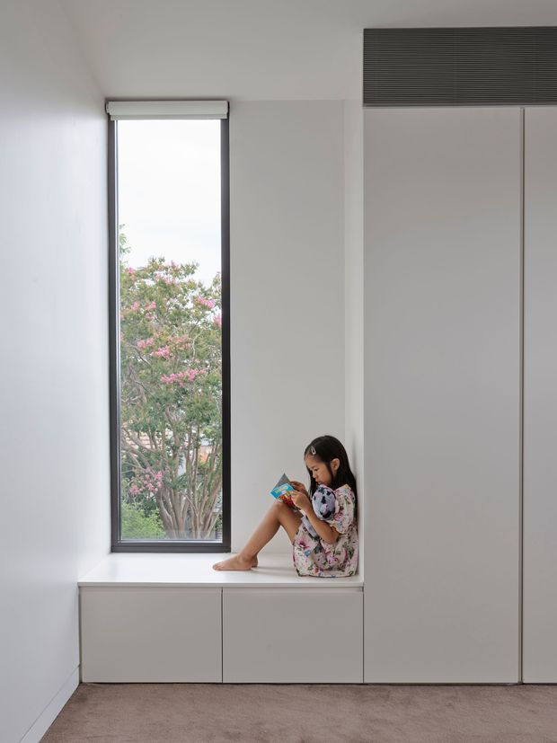 Minimal, light-filled built-in window seat and storage wall. A child sits reading on a white window bench beside a tall, narrow black-framed window. Floor-to-ceiling pale cabinetry surrounds the seat, with a dark horizontal ventilation grille above. The room has white walls, soft neutral carpeting, and a clean contemporary fitted interior.