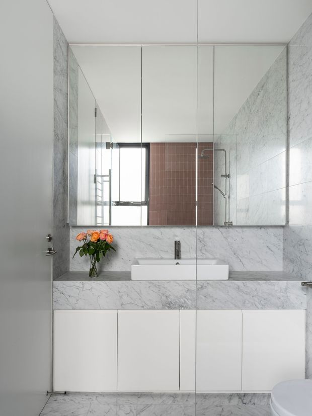 A compact contemporary bathroom with a wall-mounted white vanity, integrated basin, large mirrored wall cabinets, and a marble-clad countertop and backsplash. A glass-enclosed shower is reflected in the mirrors, featuring pale pink square wall tiles and chrome fittings. The floor and walls use white-grey veined marble, creating a bright, refined finish.