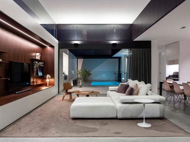A sophisticated open-plan living room with a large white sectional sofa, low round coffee tables, and a warm timber media wall with integrated shelving and television. Dark ribbed architectural screening and a partial double-height ceiling add drama, while full-height glazing or an opening connects the room to a blue-lit pool area. A dining area with a table and upholstered chairs is visible to the right. The palette is contemporary, combining white, dark charcoal, timber, and soft neutral tones.