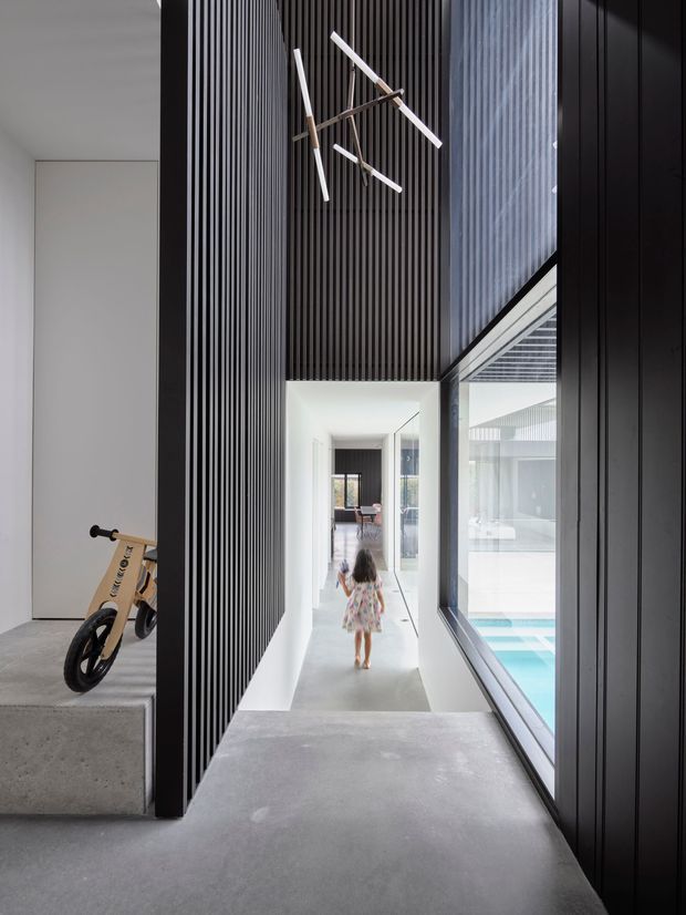 A modern interior corridor with a concrete floor and a dramatic double-height void. Black vertical slatted wall panels create a strong architectural screen, while full-height glazing on the right overlooks a bright swimming pool courtyard. A minimalist linear pendant hangs above the passage, and a child's bicycle is visible on a raised concrete platform to the left. The hallway leads toward a brighter living area beyond.