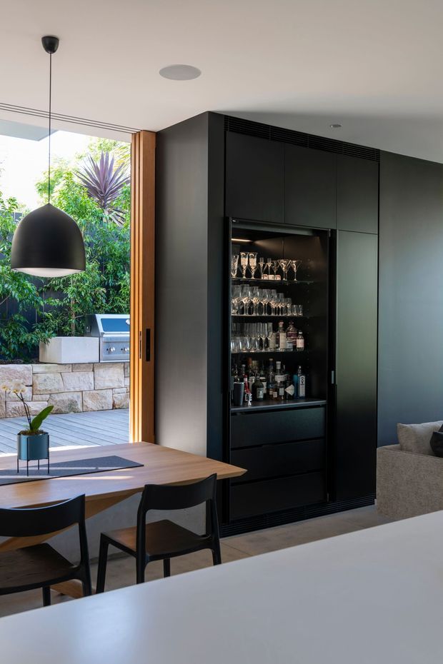 A contemporary open-plan dining and living area with a dark built-in bar cabinet displaying glassware and bottles. The room features a warm timber dining table, black dining chairs, a large black pendant light, neutral walls and cabinetry, and full-height glazed opening to a landscaped outdoor deck with a built-in barbecue and stone wall.