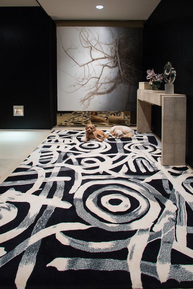 A dramatic, dark-toned interior foyer or hallway featuring a large abstract black-and-cream patterned rug, black textured wall panels, a sculptural stone console, decorative objects, and a large artwork depicting branching roots or branches. Two small dogs rest on the rug. Recessed ceiling lighting provides focused illumination.