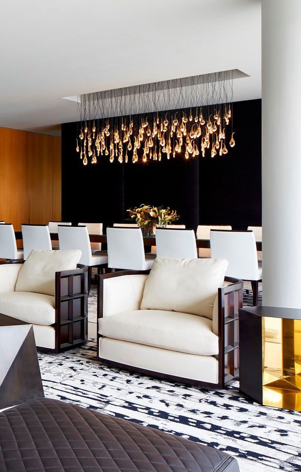 A sophisticated open-plan lounge and dining area with two cream upholstered armchairs, a dark leather ottoman, a long dining table with white chairs, patterned monochrome flooring, warm timber wall panelling, and a dramatic cluster of suspended glowing pendant lights. A black feature wall and a contemporary fireplace add contrast and luxury.