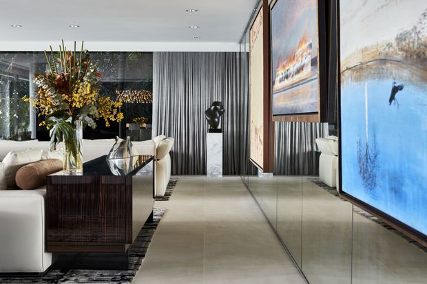 A refined contemporary living room with a neutral sectional sofa, sculptural side table, large floral arrangement, reflective mirrored wall panels, dramatic vertically ribbed dark wall treatment, and oversized artwork. Pale stone-look flooring and floor-to-ceiling glazing create a spacious, polished interior with layered reflections and gallery-like styling.
