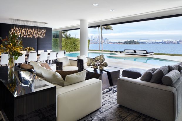 A sophisticated open-plan living and dining area with floor-to-ceiling glazing overlooking a waterfront, pool and city skyline. The room features large pale upholstered sofas, a dark glossy dining table and cabinetry, white dining chairs, a dramatic sculptural pendant installation, a dark feature wall, and polished contemporary finishes.