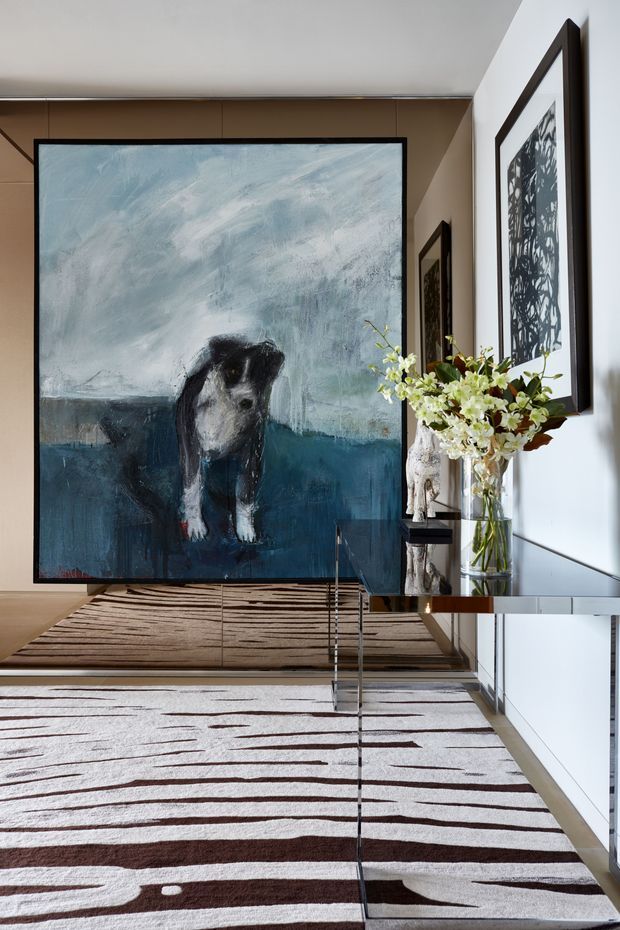 A refined contemporary hallway or entryway with a large abstract dog painting as the focal point. Warm wood wall panelling, white walls, framed monochrome artwork, a slim mirrored console table, sculptural vase arrangements, and layered neutral rugs create a sophisticated gallery-like interior.