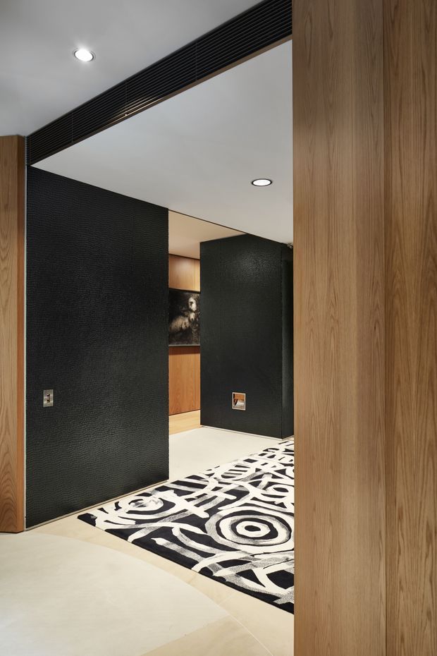 A refined interior passage or entry area framed by warm vertical timber wall panelling and dark textured feature walls. A large black-and-white graphic rug leads through the space toward built-in timber cabinetry and artwork. Recessed ceiling lighting and a long black linear vent add a contemporary architectural feel.
