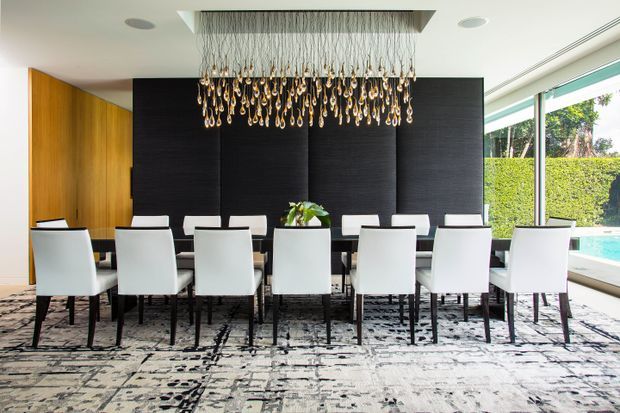 Contemporary formal dining room with a long dark dining table surrounded by white upholstered chairs. A dramatic cascading pendant chandelier hangs above, framed by a black textured feature wall and warm timber wall panelling. Floor-to-ceiling glazing opens views toward greenery and a swimming pool, while a large monochrome patterned rug anchors the room.
