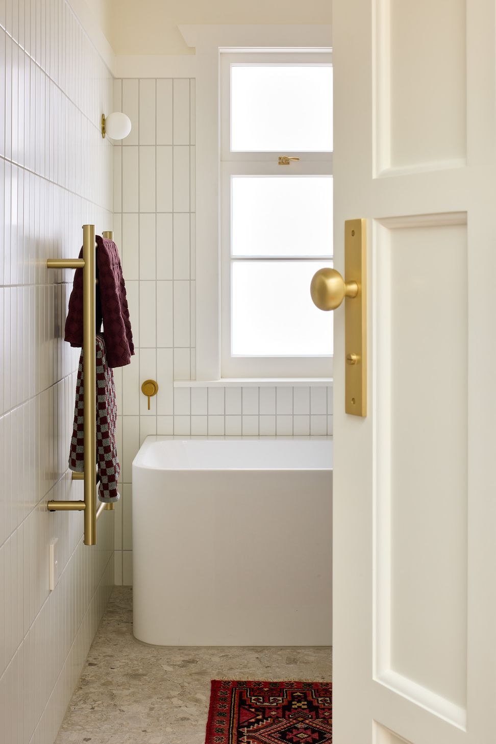 The family bathroom continues the refined material palette with terrazzo flooring, white wall tiles and brass detailing. A bath, feature wall light and restored traditional door fitted with Iver hardware celebrate the home's blend of old and new.
