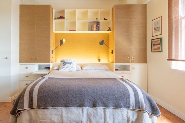 A warm, compact bedroom with a built-in wall-to-wall storage and bedhead unit. The room features light wood cabinetry, open shelving with books and decor, integrated bedside drawers, wall-mounted reading lamps, and a neatly made bed with a grey and white striped throw. A timber floor, framed artwork, and a timber venetian blind complete the bright, contemporary interior.