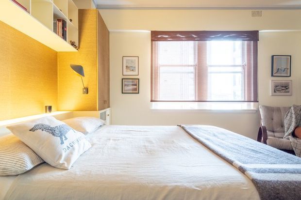 Bright, warm bedroom with a large bed, white bedding, a mustard-yellow textured built-in headboard and wall panel, integrated bedside lighting, overhead shelving and cabinetry, and a broad window with blinds. A reading chair and framed artwork sit to the side, creating a cozy, cheerful retreat.