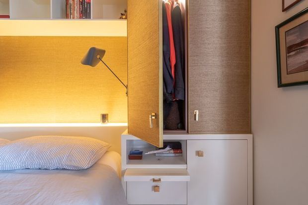 Compact, warm-toned bedroom with a built-in timber feature wall, integrated reading light, open shelving, and a fitted wardrobe with drawers and concealed clothing storage. The bed occupies the lower-left portion, while framed artwork and a neutral painted wall appear on the right.