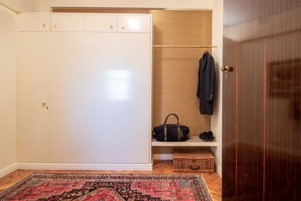 A compact entryway with built-in cream cabinetry, an open coat and shoe nook, a hanging rail, wicker storage basket, and patterned area rug. Herringbone timber flooring and a dark wood door add warmth and contrast.