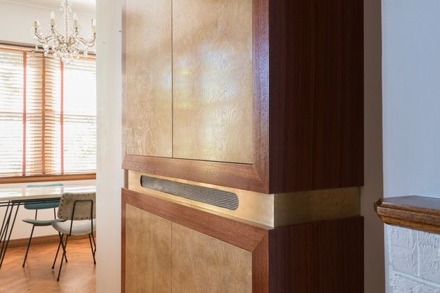 Close-up interior view of a custom timber built-in cabinet or wall-mounted joinery unit, featuring warm figured wood panels, dark timber side panels, horizontal trim, and a recessed slatted detail. A bright dining area with timber flooring, a chandelier, and timber venetian blinds is visible to the left, while a painted brick fireplace or wall edge appears at right.