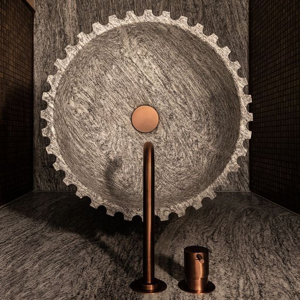 Dramatic close-up of a sculptural bathroom vanity installation featuring a large circular, gear-edged stone basin or wall feature, a central copper-toned faucet, and a matching cylindrical fixture. Dark textured stone surfaces and black tiled side walls create a moody, luxurious setting.