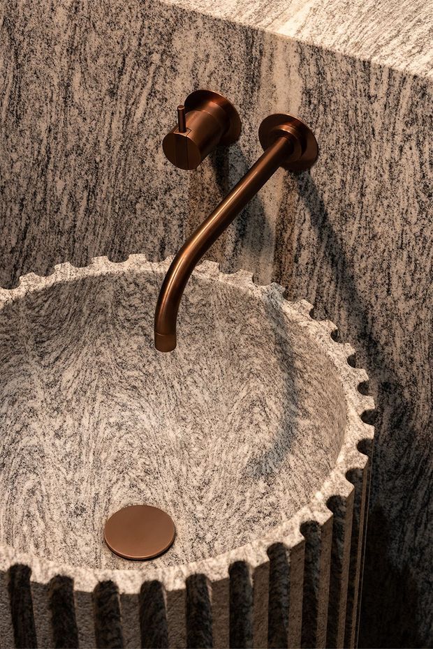 Close-up of a sculptural bathroom basin carved from richly veined grey stone, with a fluted scalloped rim and matching stone wall surfaces. A wall-mounted curved faucet and circular drain are finished in warm bronze, creating a refined, tactile and highly detailed bathroom installation.
