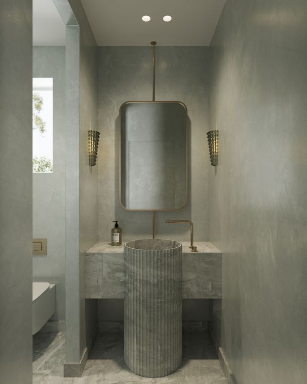 A compact, refined powder room with a sculptural fluted pedestal basin centered beneath a tall rounded-rectangle mirror. The room features grey-green textured wall finishes, matching stone-look flooring and vanity cabinetry, a pale stone benchtop, warm brass-framed mirror and tapware, and decorative wall sconces. A toilet is partially visible through the opening on the left. The mood is subdued, luxurious, and cohesive, with carefully integrated lighting and materials.