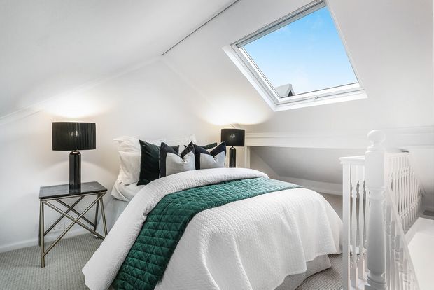 Bright attic-style bedroom with a sloped white ceiling, a large skylight, and a neatly made bed layered in white bedding with a dark teal quilted runner and pillows. Black bedside lamps and a slim metallic side table add contrast, while a white stair balustrade is visible in the foreground.