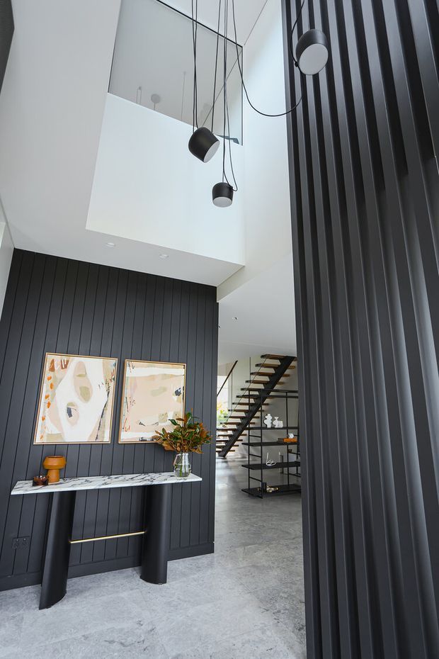 A dramatic contemporary residential entryway with a double-height void and floating pendant lights. The space features dark vertical wall panelling, a matching fluted screen, a slim marble console with black supports, abstract artwork, polished grey concrete flooring, and a view toward an open-riser staircase and shelving.