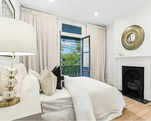 Bright, refined bedroom with a white upholstered bed dressed in layered white bedding and neutral cushions. Full-height cream curtains frame an open glazed door leading to a small balcony, while a traditional black fireplace with a white mantel and round metallic mirror provide architectural character. Pale timber floorboards and recessed ceiling lights complete the calm, elegant interior.