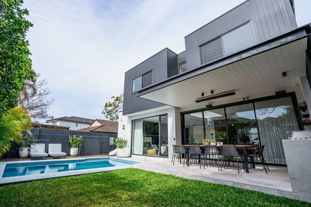 Contemporary two-storey residential home viewed from the backyard, featuring dark grey vertical cladding, expansive sliding glass doors, a covered outdoor dining terrace, and a rectangular swimming pool beside a manicured lawn. The clean-lined design includes a projecting upper level and a light-coloured soffit with recessed lighting.