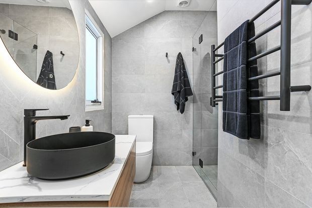 A contemporary bathroom with large-format grey tiled walls and floor, a floating timber vanity with a white stone benchtop, matte black vessel basin and tapware, a frameless glass shower, wall-hung toilet, round backlit mirror, and black heated towel rail. The space is compact, bright, and refined with strong monochrome contrast.