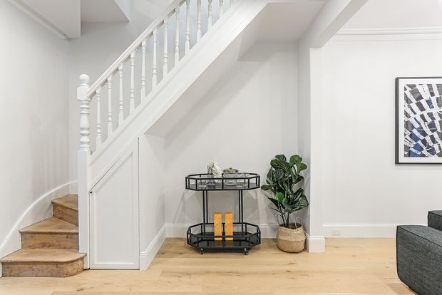 Bright white-painted stairway and hallway area with traditional turned balusters, timber stair treads, decorative wall mouldings, and pale timber flooring. A black metal bar cart, potted plant, framed artwork, and part of a dark sofa add furnishings to the space beneath and beside the staircase.