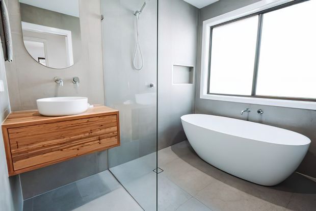 Contemporary bathroom with a freestanding white oval bathtub beside a large window, a frameless glass shower enclosure, wall-mounted timber vanity with white vessel basin, and grey tiled wall and floor finishes. The space is bright and minimal with warm timber joinery providing contrast.