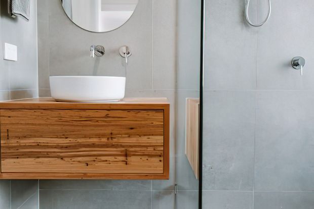 Contemporary bathroom featuring a wall-mounted timber vanity with a white vessel basin, minimalist wall-mounted chrome tapware, large pale grey wall tiles, and a frameless glass shower enclosure. An oval mirror and simple white switch contribute to the clean, modern fit-out.
