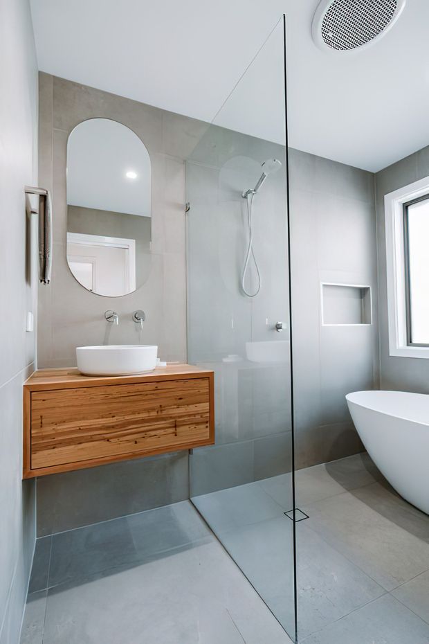 A bright, contemporary bathroom with large grey wall and floor tiles, a frameless glass shower enclosure, a wall-mounted timber vanity with a white vessel basin, an arched mirror, recessed shower niche, freestanding white bathtub, and a window providing natural light.