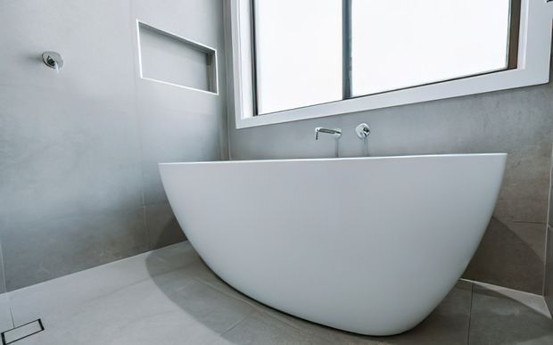 Contemporary bathroom featuring a freestanding white oval bathtub positioned beneath a large window, with wall-mounted chrome tapware and a recessed wall niche. The room has large grey stone-look wall and floor tiles, a clean minimal fit-out, and abundant natural light.